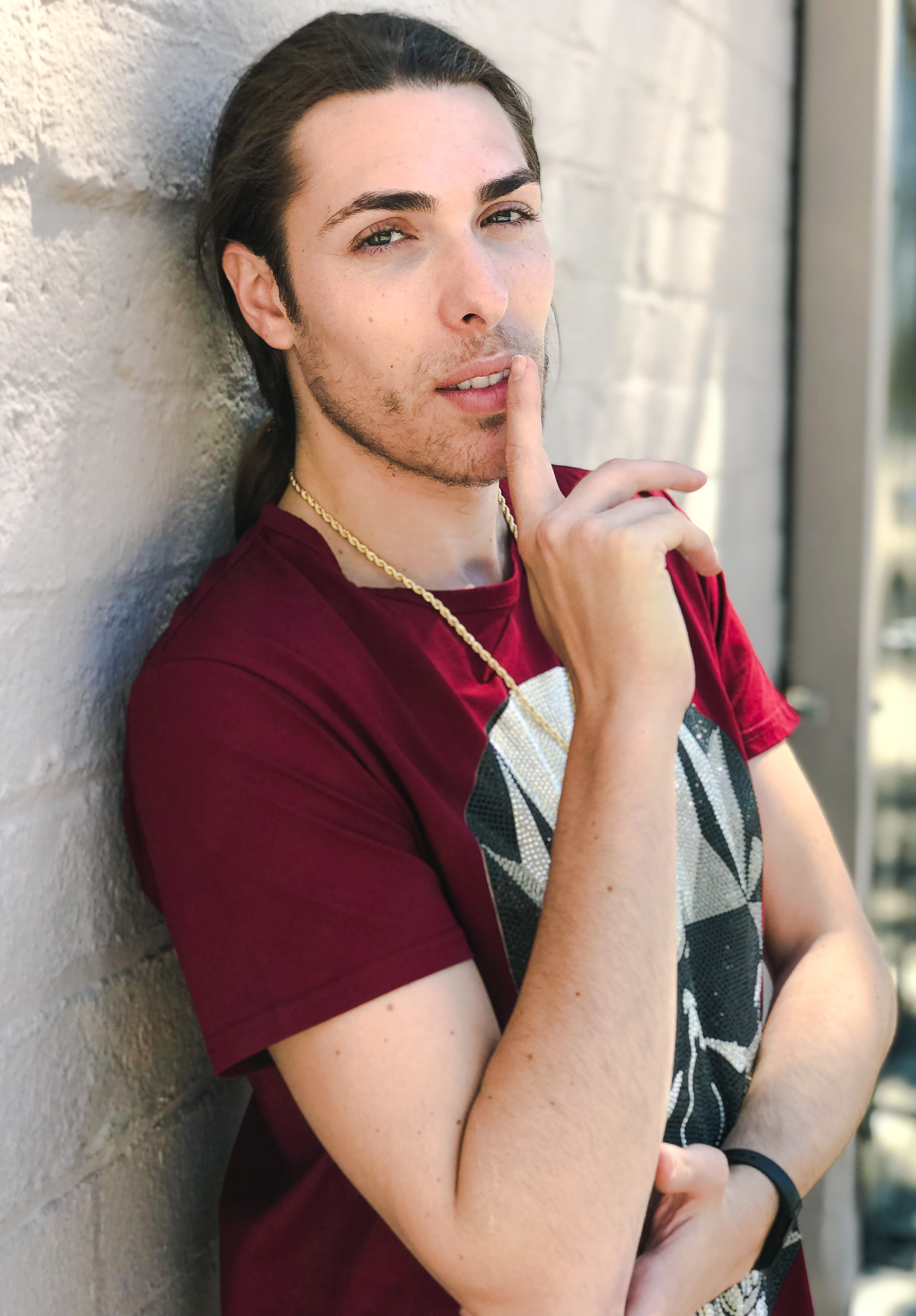 Alex Veach (Alexander Veach) LA streetwear, burgundy tee, finger-to-lips gesture on white wall.