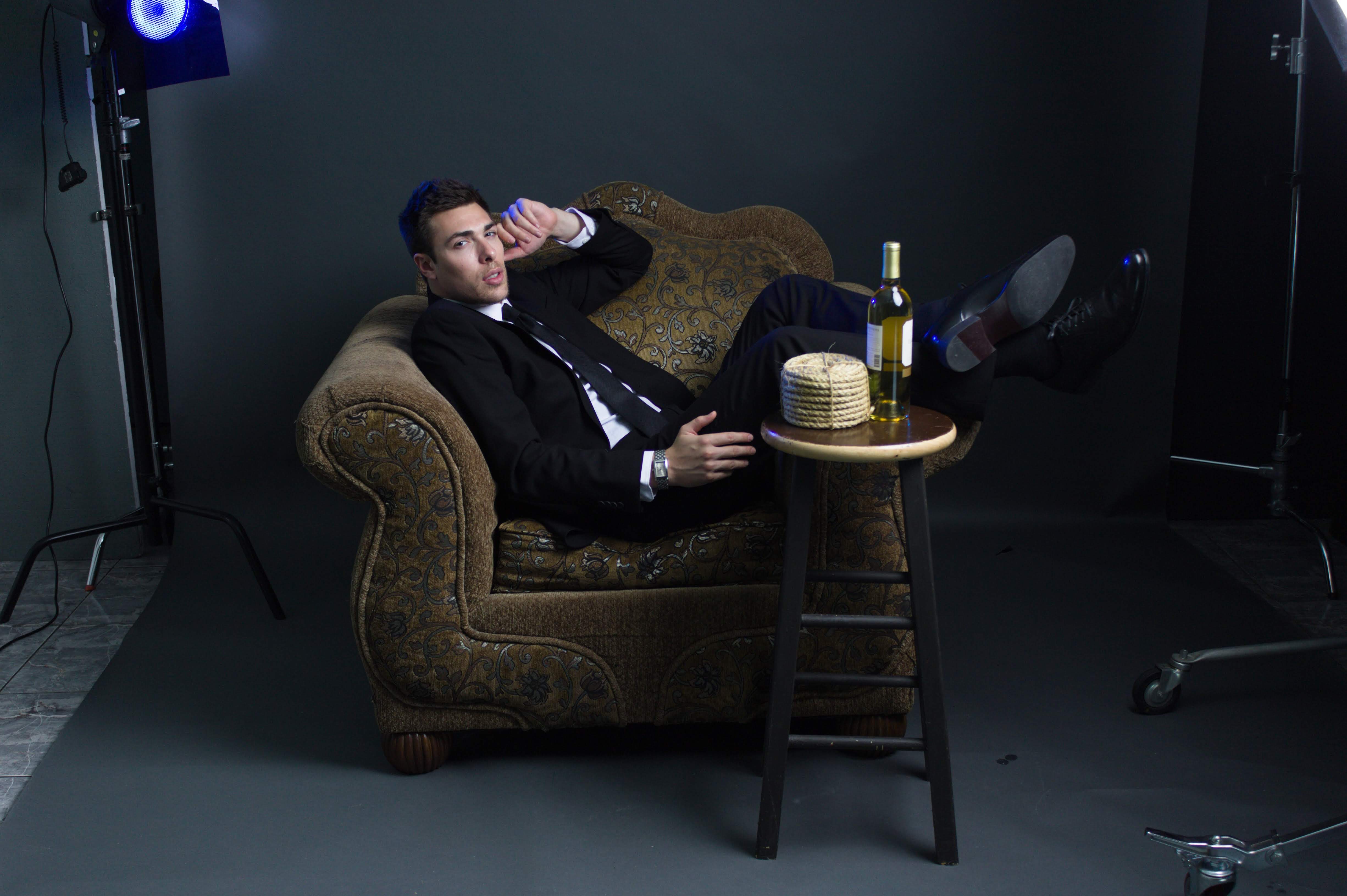 Alex Veach (Alexander Veach) black-suit editorial, reclined on armchair with full studio scene visible.